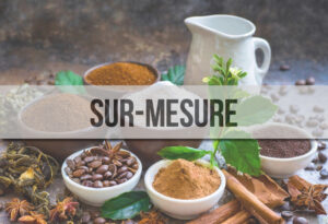 Sur une table, plusieurs petits bols contenant des grains de café, de la poudre de lait, de chicorée, des infusions, du thé, des épices, des herbes aromatiques et un pichet de lait. Et un bandeau au premier plan où il est écrit "sur-mesure".